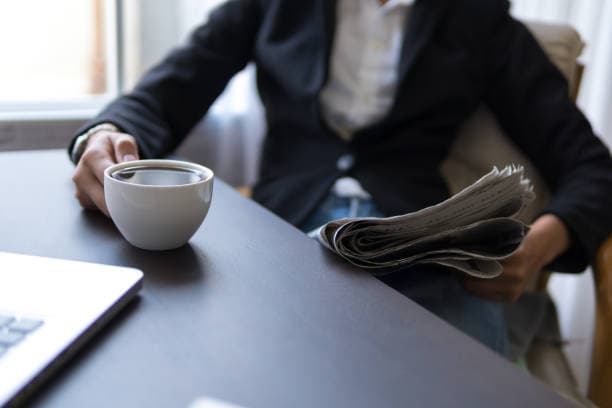 Person in a blazer holding a cup of coffee and newspaper at a desk.